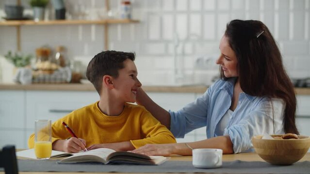 A smiling boy sits at the table with his mother, who helps him do his homework. A long-haired brunette woman kisses her son on the forehead. Generative AI