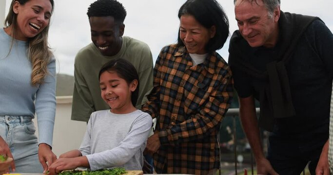 Multiracial friends having fun preparing food with girl during weekend day - Multi generational people doing barbecue at home's rooftop - Latin child cutting herbs