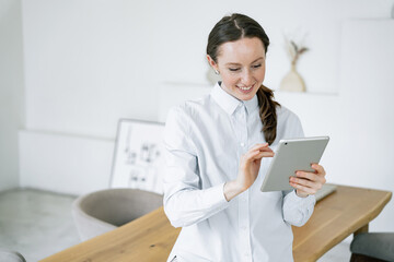 A creative freelancer in a blue shirt is immersed in work, using a tablet in a bright office. Daily work online chat in social networks.