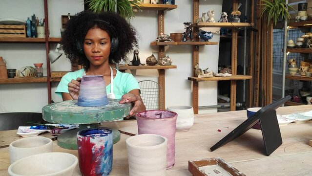 Crafting Beauty: A focused artisan, wearing headphones, molds a ceramic pot on a potter's wheel, surrounded by shelves of finished works, embracing the creative process.