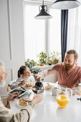 Gay man pouring coffee near partner and daughter during breakfast at home. 