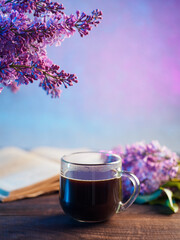 A cup of coffee on a wooden table, a bouquet of lilacs and an open book, copy space
