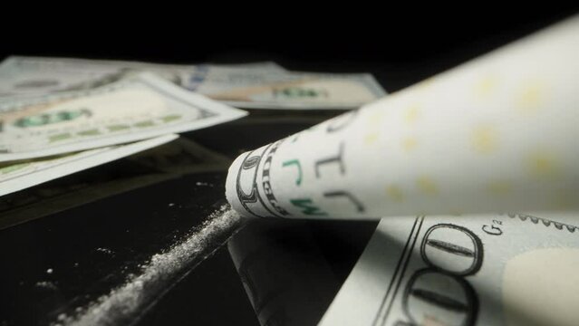 Imitation of cocaine use through a folded one hundred dollar bill, banknotes are scattered on a black table. Dolly slider extreme close-up.