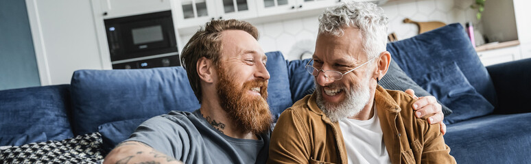 Tattooed gay man hugging cheerful mature partner in living room, banner. 