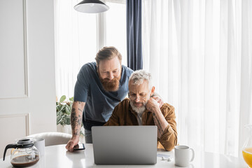 Tattooed gay man hugging mature partner using laptop near coffee at home. 