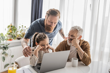 Homosexual fathers using laptop with carefree kid during online education near drinks at home. 