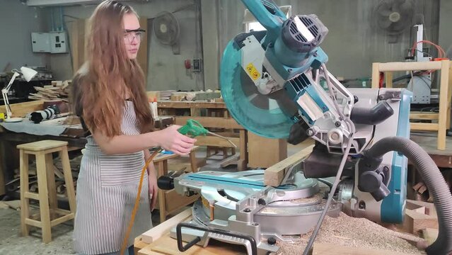 Craftswoman at Work: A young woman in protective eyewear and an apron operates a miter saw in a workshop, carefully preparing to cut a piece of wood.