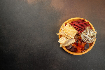Naklejka premium snacks for beer, Dried fish mix on wood plate. dried fish chorizo sausage meat carpaccio on a dark background. place for text, top view
