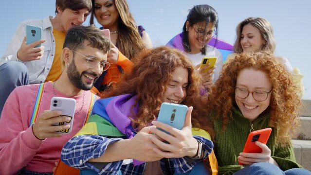 Group young friends watching mobile phone and celebrating gay pride festival day together. Cheerful LGBTIQ community outdoors checking social networks. Lesbian, gay, transgender