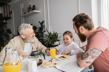 Positive girl writing on notebook near homosexual parents and pancakes at home.