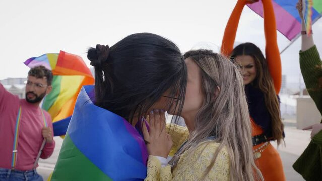 Two young loving girls kissing on gay pride festival day outdoors. Group of friends celebrating lgbt party on background with rainbow flag and fans. Generation z and types of sexuality.