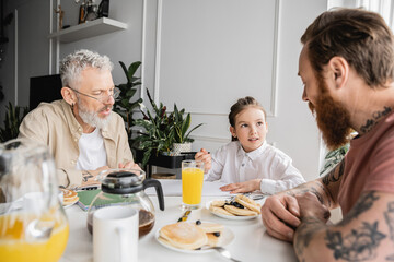 Preteen girl talking to homosexual fathers near notebook and pancakes at home.