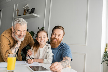 Smiling gay parents with daughter looking at camera while doing homework at home. 