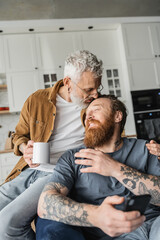 Homosexual man holding coffee and kissing tattooed partner with smartphone at home. 