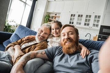 Smiling preteen kid hugging same sex parents and looking at camera on couch at home. 