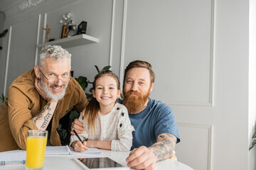 Smiling gay parents and daughter looking at camera near notebook and digital tablet at home. 