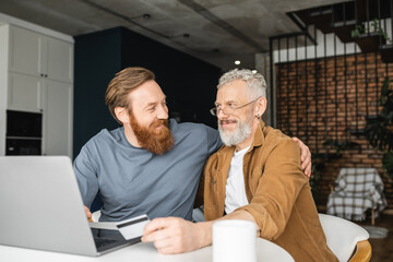 Positive gay man hugging partner with credit card near laptop and coffee at home. 