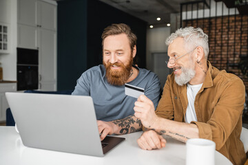 Fototapeta premium Carefree gay man holding credit card near partner with laptop at home. 