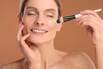 Woman applying foundation on face with brush against brown background