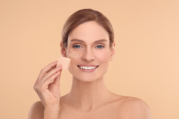 Woman blending foundation on face with makeup sponge against beige background