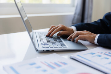 A business man working on a laptop comparing details from graphs Business income chart in real estate, tax, management, marketing, technology in private office.