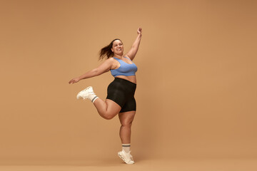 Fototapeta na wymiar Young, active, smiling, overweight woman training in sportswear against brown studio background. Workout with positivity. Concept of sport, body-positivity, weight loss, body and health care