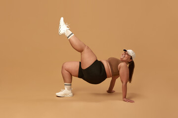 Positive, young, overweight woman training in stylish and comfortable sportswear over brown studio background. Full-body workout. Concept of sport, body-positivity, weight loss, body and health care - obrazy, fototapety, plakaty