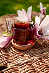Hot tea on the background of a wicker basket with magnolia flowers. Spring still life composition. Good morning concept. Transparent cup with tea on the terrace