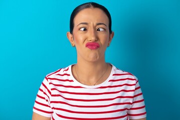 beautiful woman wearing striped T-shirt over blue studio background crosses eyes, puts lips, makes...