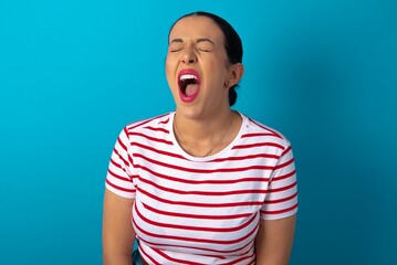 Stressful beautiful woman wearing striped T-shirt over blue studio background screams in panic, closes eyes in terror, keeps hands on head, finds out terrified news, can't believe it.