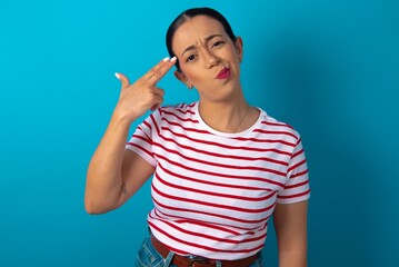 Fototapeta premium Unhappy beautiful woman wearing striped T-shirt over blue studio background imitates gun shoot makes suicide gesture keeps two fingers on temples.