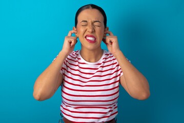 Stop making this annoying sound! Unhappy stressed out beautiful woman wearing striped T-shirt over blue studio background making worry face, plugging ears with fingers, irritated with loud noise.