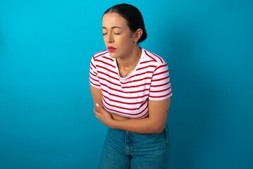 beautiful woman wearing striped T-shirt over blue studio background suffering from strong stomachache.