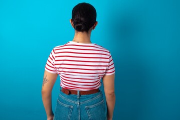beautiful woman wearing striped T-shirt over blue studio background standing backwards looking away with arms on body.