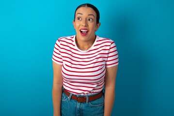 Surprised beautiful woman wearing striped T-shirt over blue studio background, shrugs shoulders, looking sideways, being happy and excited. Sudden reactions concept.