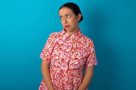 Dissatisfied beautiful woman wearing floral dress over blue studio background purses lips and has unhappy expression looks away stands offended. Depressed frustrated model.