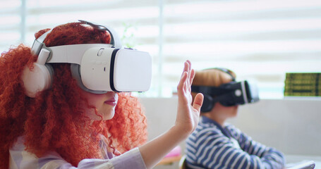 Amazed preteen girl using VR headset while sitting with classmates at classroom. Excited students swiping on invisible screen and listening to teacher during lesson.