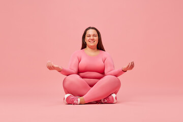 Young, smiling, overweight woman training in sportswear, sitting in yoga lotus pose, meditating against pink studio background. Concept of sport, body-positivity, weight loss, body and health care