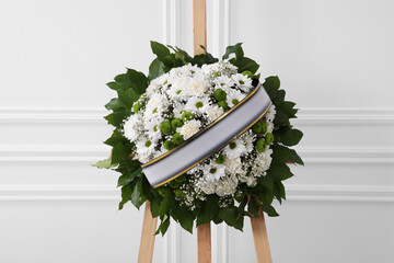 Funeral wreath of flowers with ribbon on wooden stand near white wall indoors © New Africa