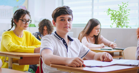 Portrait of nice Caucasian teen clever schoolboy in glasses sitting at desk in classroom with laptop computer and looking at camera. Among kids. Indoors. Smart boy learning in school concept.