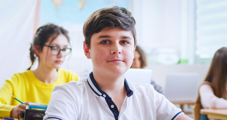 Portrait of nice happy Caucasian teen clever schoolboy sitting at desk in classroom and smiling to camera. Among kids. At school. Little teenage boy learning in school concept. Close up.