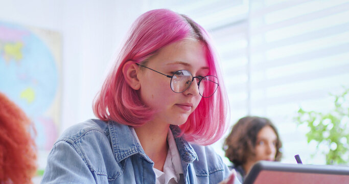 Pensive schoolgirl using digital tablet while classmates studying on background at classroom. Focused female pupil with pink hair browsing internet on pad during lesson.