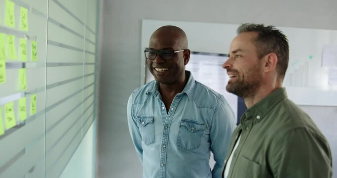 Two diverse businessmen laughing during a brainstorming session together with adhesive notes on a glass wall of an office