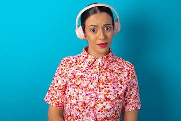 Serious displeased beautiful woman wearing floral dress over blue studio background looks puzzled at camera being angry wears stereo headphones listens music while walking at street