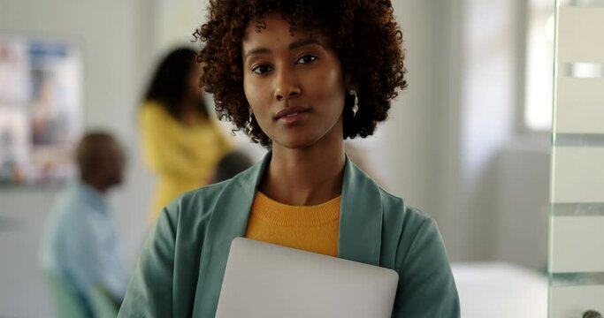 Confident young businesswoman holding a laptop standing at the door to a boardroom with coworkers meeting in the background