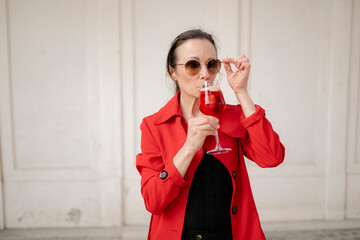 Frau, 40+, mit Sonnenbrille im roten Mantel mit einem Glas Spritz