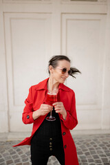 Frau, 40+, mit Sonnenbrille im roten Mantel mit einem Glas Spritz