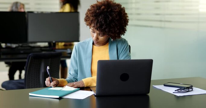 Young businesswoman making notes and using a laptop at her desk in an office with female colleagues talking in the background