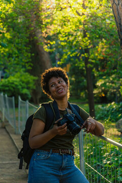 Happy Young Black African American Woman Photojournalist With Dark Curly Hair Laughing Happily Looking At Friend With Photo Camera Hanging From Neck In Green Park At Sunset With Sun Behind Backlightin