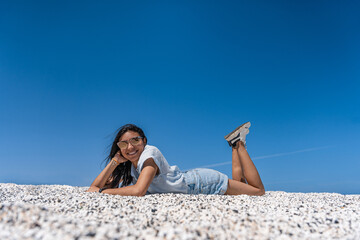 mujer latina acostada boca abajo en arena blanca con cielo azul de fondo con una sonrisa y pose que...